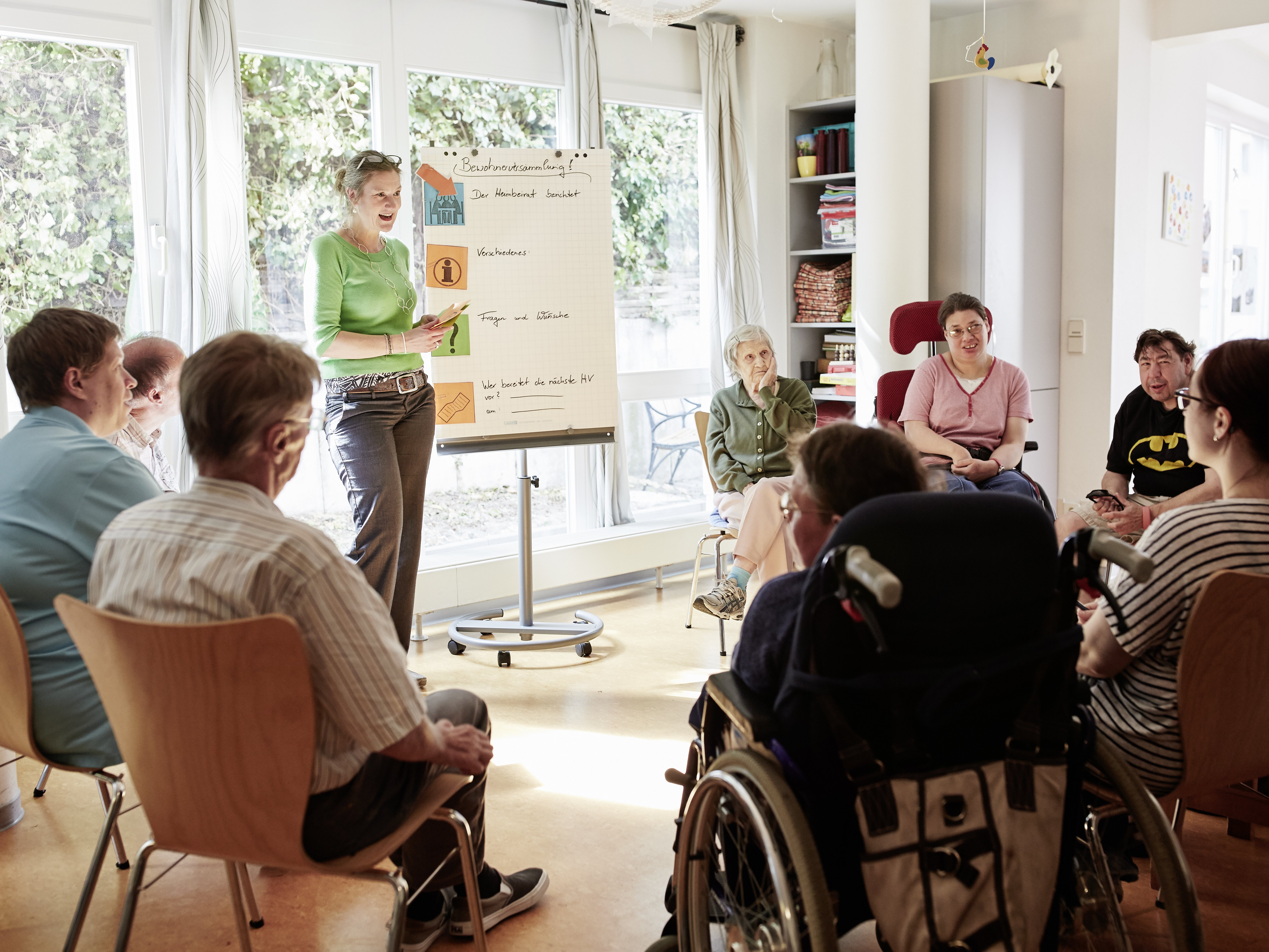 Foto von einer Schulung für Menschen mit und ohne Behinderung. Die Zuhörer sitzen in einem Stuhlkreis. Vorne steht eine Referentin und zeigt etwas auf einem Flipchart.