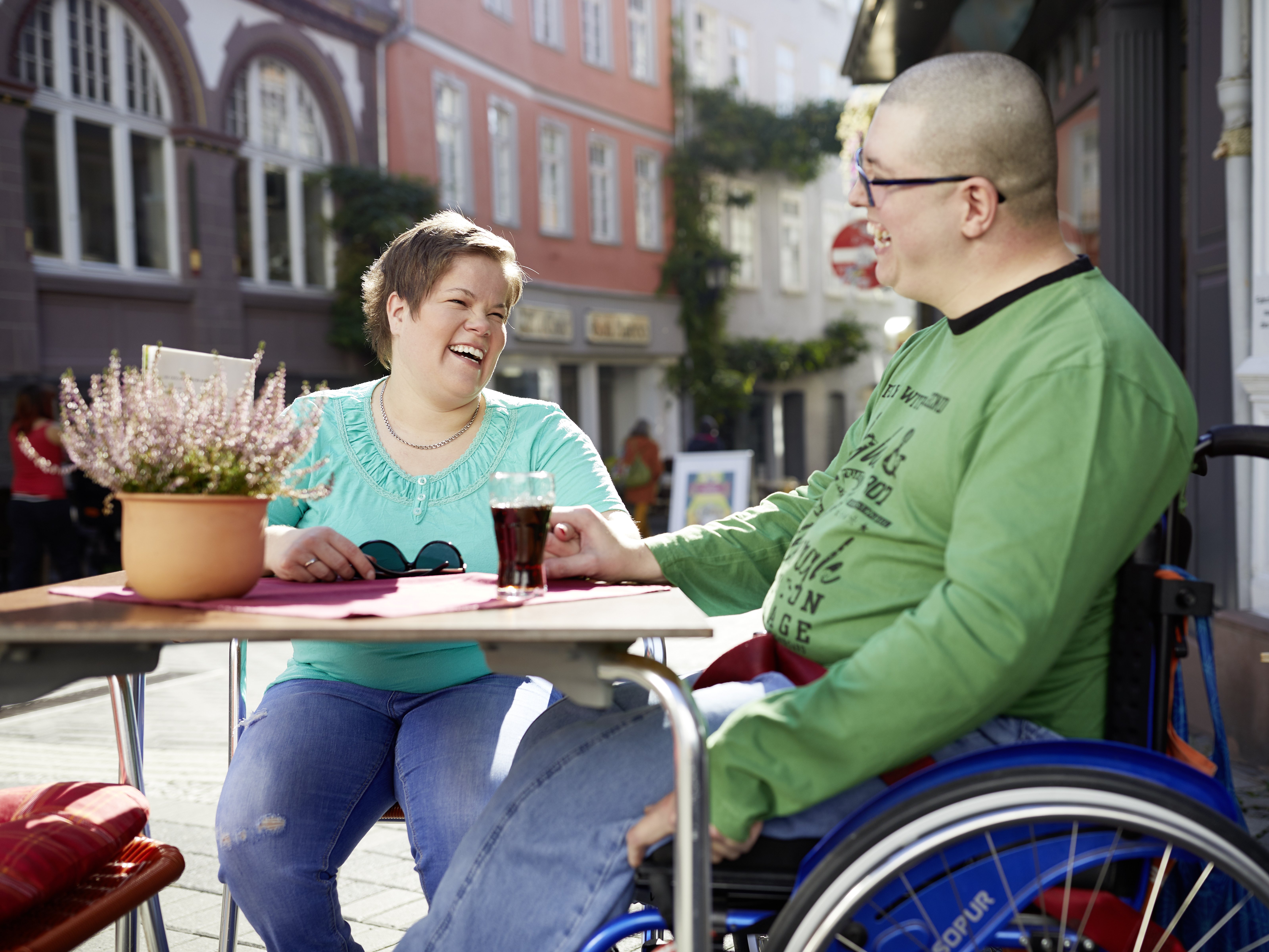 Eine Frau sitzt zusammen mit einem Mann im Rollstuhl am Tisch eines Straßencafes.