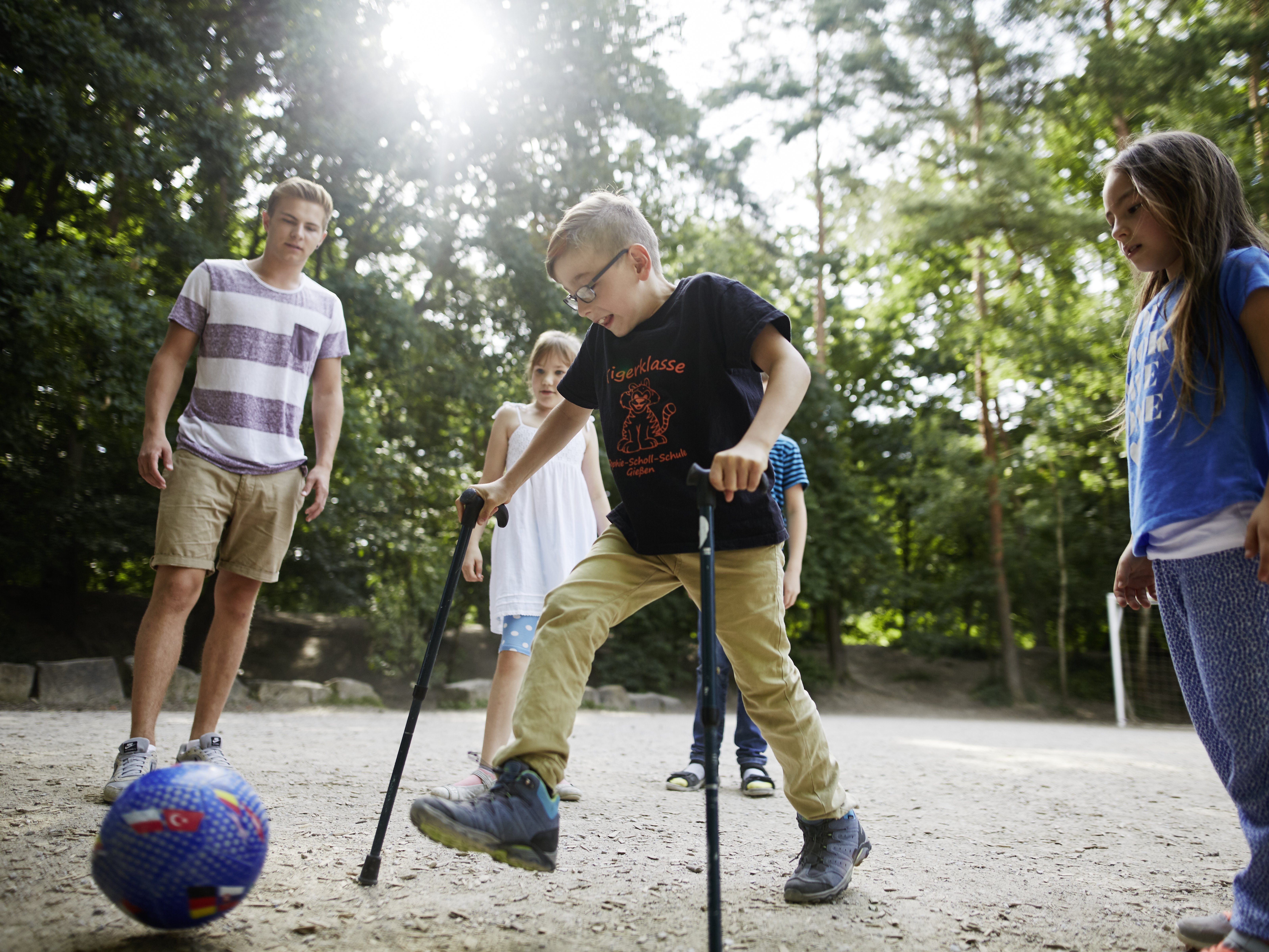 Foto von Kindern auf einem Spielplatz, darunter ein Junge mit Krücken der einen Ball kickt. Dabei ein junger Mann als Betreuer der Lebenshilfe.