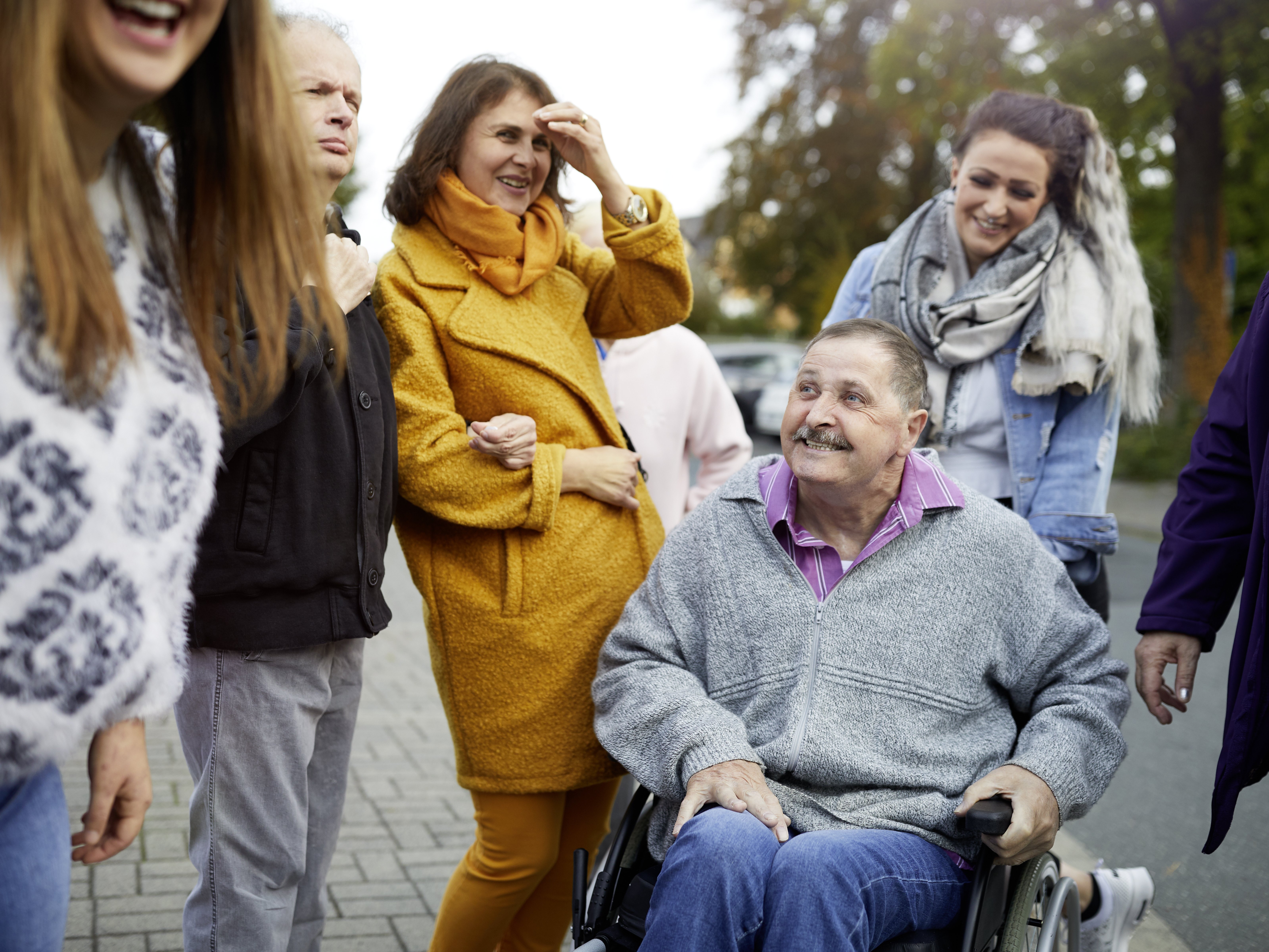 Foto einer Gruppe von erwachsenen Menschen mit Behinderung und deren Betreuern. Sie sind im Freien unterwegs. Ein Mann wird im Rollstuhl geschoben.