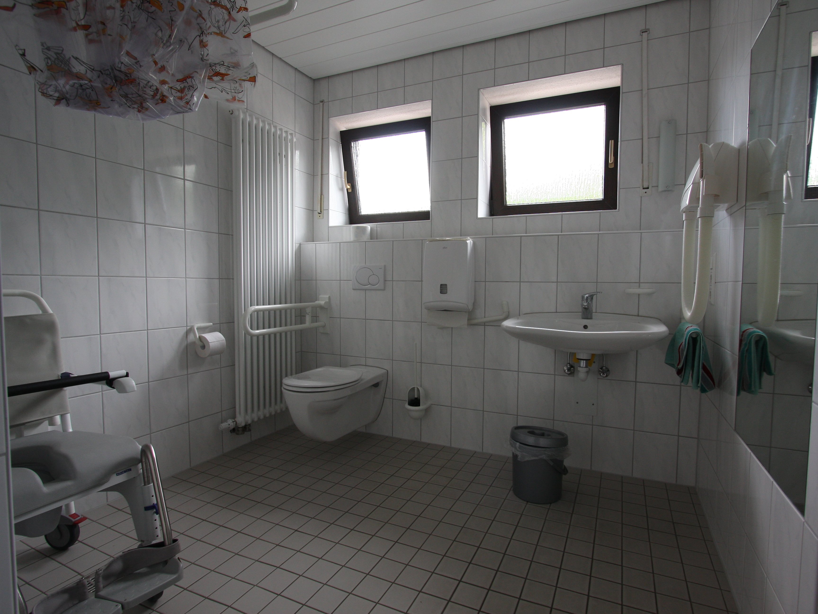 Foto von einem barrierefrei ausgestatteten Bad mit rollstuhlgerechter Dusche mit Duschstuhl, WC mit Haltegriffen und Waschbecken.